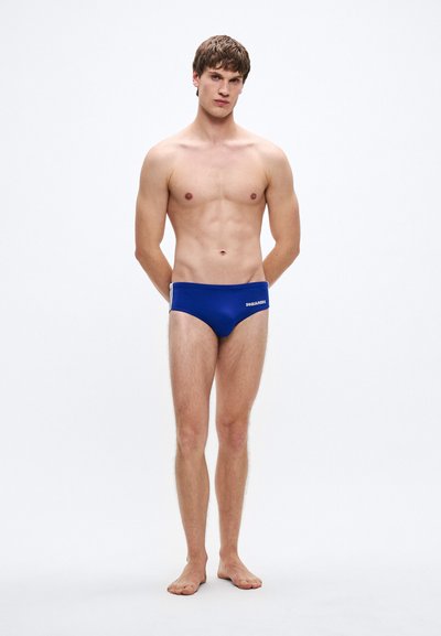 Blaue Badehose mit einfarbigem Design und weißem Logo, mit elastischem Bund. Das Modell steht mit hinter dem Rücken verschränkten Armen.