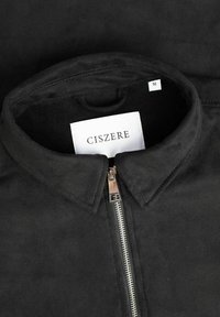 Giacca in suede nera con chiusura a zip, colletto e un'etichetta con la scritta "CISZERE" di taglia "M". Texture liscia con accenti minimi.