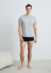 T-shirt en coton gris à manches courtes, associé à des boxers noirs. Le mannequin se tient pieds nus sur un tapis clair, avec un canapé doux à proximité.