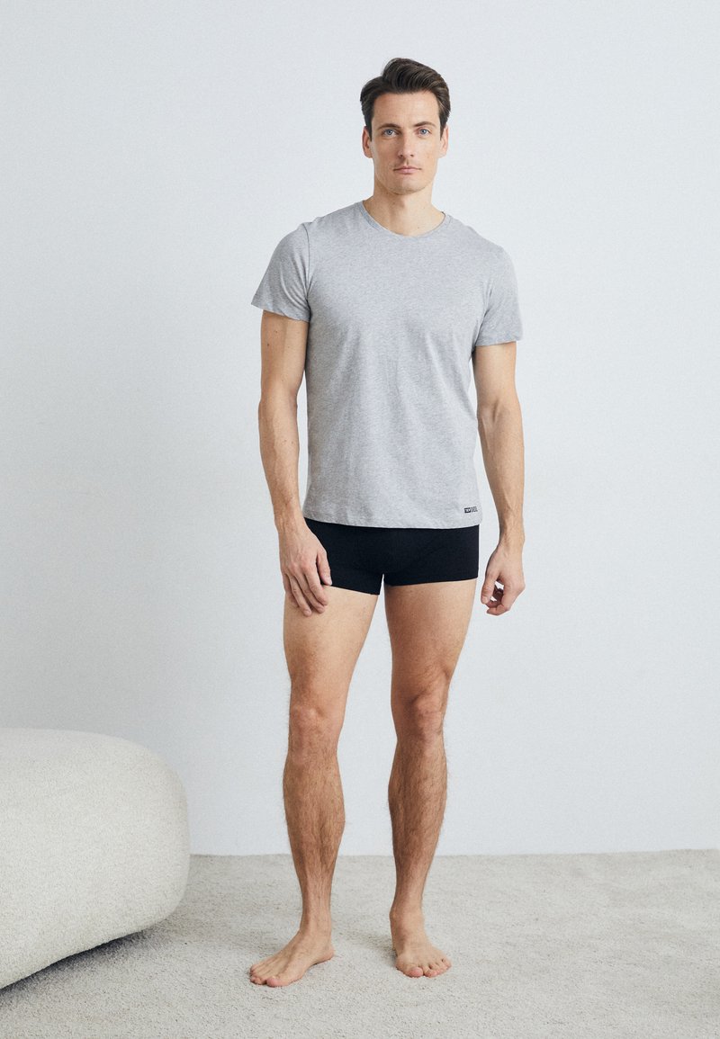 T-shirt en coton gris à manches courtes, associé à des boxers noirs. Le mannequin se tient pieds nus sur un tapis clair, avec un canapé doux à proximité.