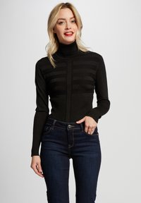 Zwarte coltrui met ribbeltextuur en horizontaal gestreept patroon; gecombineerd met donkere spijkerbroek. Model staat tegen een effen achtergrond.