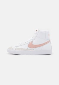 Vita högstukna sneakers med en rosa Nike swoosh och velur tåkappa. Har vita snören och en struktur i ovandelen. Accentuerad med logotypdetalj på hälen.