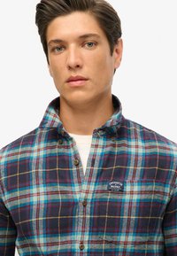 Camicia in flanella a quadri blu e navy con un sottile logo ricamato, colletto con bottoni e bottoni visibili. Il tessuto appare morbido e strutturato.