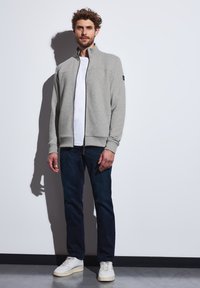 Veste zippée gris clair avec un motif texturé, des accents noirs et un col haut. Portée sur un t-shirt blanc et associée à un jean foncé et des baskets blanches.