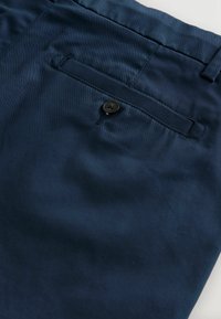 Tengerészkék szövet közelről, nadrág hátsó zsebe egy fekete gombbal és övbújtató részlettel.