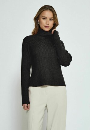 Black ribbed turtleneck sweater with a relaxed fit and long sleeves, paired with light-colored trousers, against a plain backdrop.