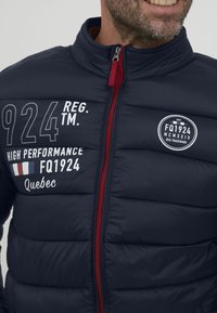 Marineblå puffjakke med rød glidelås, med broderte tekster: "924 REG. TM," "HIGH PERFORMANCE FQ1924," og "Quebec." Teksten inkluderer fransk flagg.