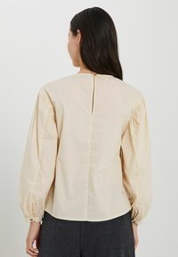 Kahvapehme blouse puffed sleeve'idega, seljal nööbiga kinnitamine ja kogutud mansetid. Sile kangas ja vaba lõige, lõpeb vöökohas.
