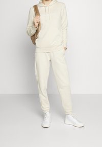 Sweat à capuche beige avec une poche kangourou à l'avant et un logo texte. Joggeurs beige assortis et baskets blanches complètent la tenue. Sac marron porté en bandoulière.