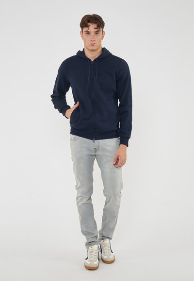 Marinblå zip-up hoodie med snörning i huvan, sidofickor och ribbade muddsydd, parat med ljusgrå jeans och vita sneakers.