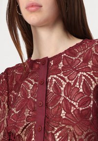 Burgundy lace top featuring floral embroidery, a round neckline, and six front buttons. Sheer texture with detailed patterns.