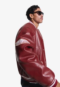 Tânăr purtând ochelari de soare închiși la culoare și o jachetă varsity roșie din piele cu patch-ul „World Leisure” pe mânecă, cu fața spre dreapta.