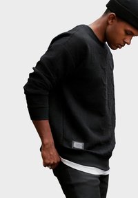 Felpa nera con texture in rilievo, polsini a costine e una patch logo rettangolare all'orlo, indossata sopra una maglietta bianca.