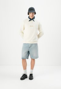 Cremet sweatshirt med broderet tekst, lyse denimshorts, grå sokker og sorte sko; har mørk kasket og mønstret bandana.