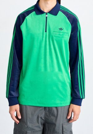 Polo vert à manches longues avec une fermeture éclair partielle, manches bleu marine et trois rayures vertes. Comprend une poche poitrine et un logo brodé sur la gauche.