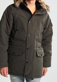 Uomo che indossa un parka invernale verde oliva con cappuccio foderato in pelliccia, chiusura a bottoni e cerniera, e tasche frontali con patta, abbinato a jeans azzurri chiari.