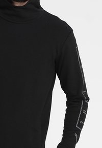 Jersey negro de manga larga con cuello alto, confeccionado en algodón, con un diseño ajustado, text en gris a lo largo de la manga izquierda y una textura suave.