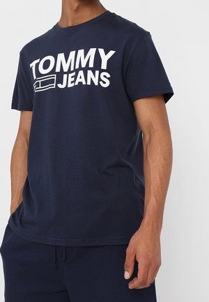 Man wearing navy blue short-sleeve T-shirt with large white "TOMMY JEANS" text and logo, paired with navy blue pants.