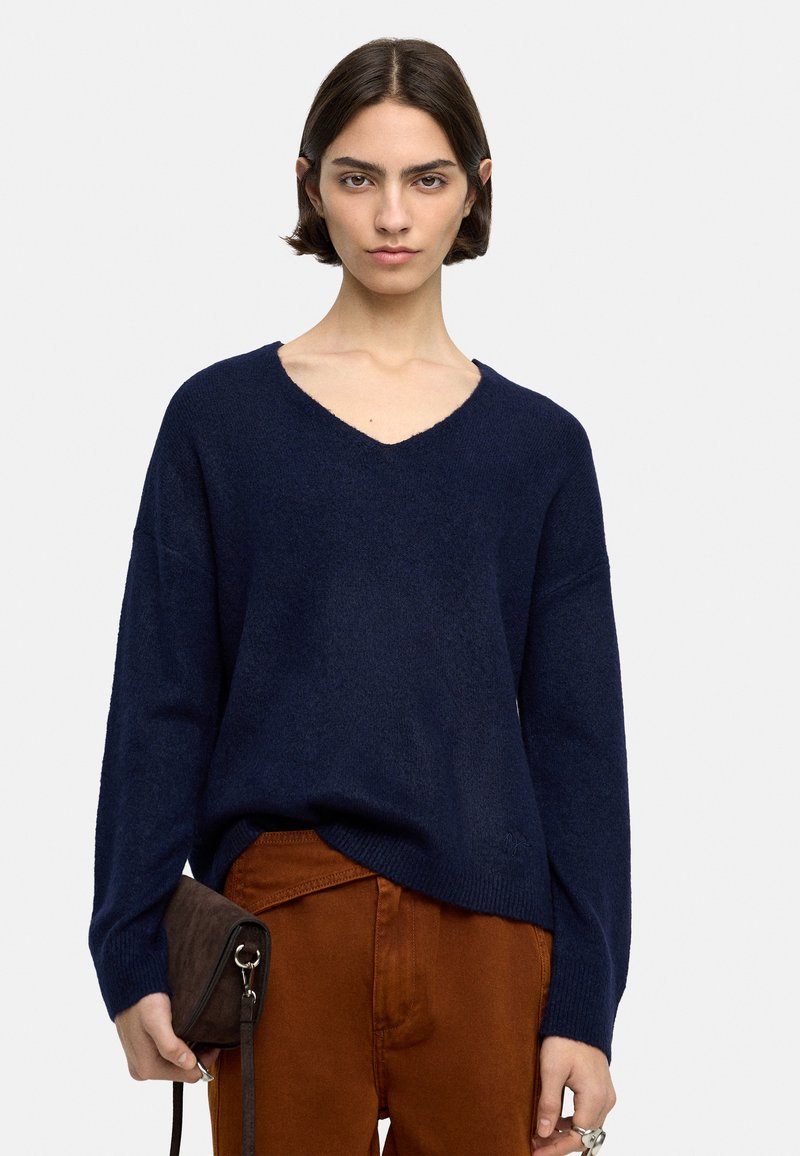 Granatowy sweter typu v-neck z długimi rękawami, wykonany z teksturowanej dzianiny. Połączony z spodniami w kolorze rdzy i małą, ciemnobrązową torebką.