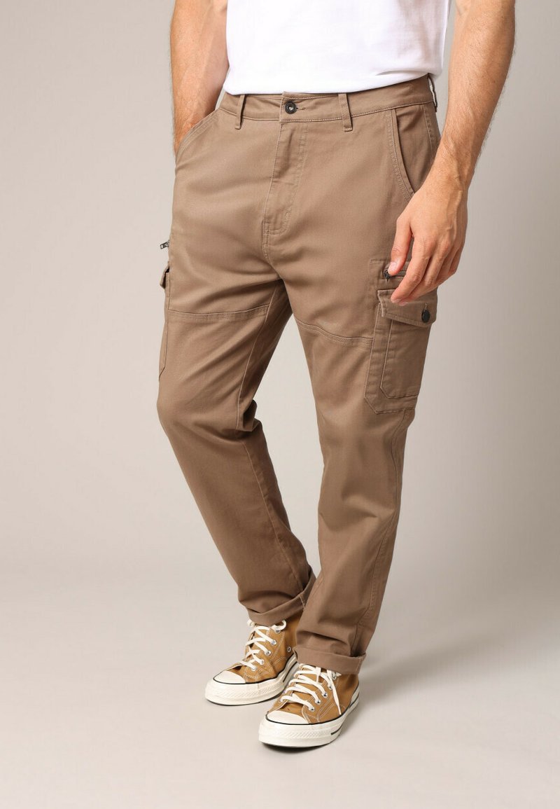 Tanskleurige cargobroek van een rechte snit, met meerdere zakken en een knoopsluiting. Draagt bij bruine sneakers en een wit shirt.