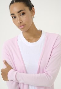 Cardigan rosa chiaro realizzato in morbido tessuto a maglia, con design aperto, caratterizzato da maniche lunghe e polsini a coste, indossato sopra una t-shirt bianca.