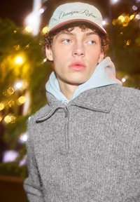 Grau gestrickter Pullover mit hohem Kragen und Reißverschluss, darüber getragen ein hellblauer Hoodie. Kombiniert mit einer blassen Mütze mit Schriftzug.