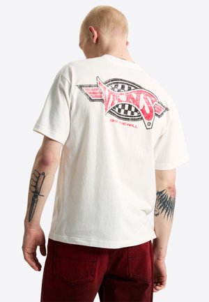 Hombre con tatuajes que lleva una camiseta Vans blanca con un logo rojo y negro y pantalones rojos, visto desde atrás.
