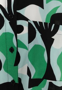 Chemise à manches courtes en tissu bleu clair, vert et noir ornée d'un motif abstrait audacieux, avec des boutons noirs et une poche plaquée.