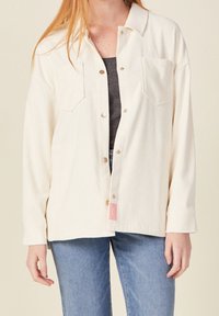 Cream corduroy shirt jacket with a button-up front, two chest pockets, and rolled sleeves. Features a small pink tag at the hem.