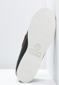 Chaussure en cuir noir avec des accents en cuir marron, arborant une semelle en caoutchouc blanche. La semelle présente un motif de sculpture et un logo Timberland.