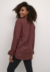 Camicia bordeaux in tessuto liscio con piega posteriore, maniche lunghe e polsini con bottoni, abbinata a jeans scuri su uno sfondo neutro.