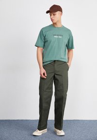 Mėlynos spalvos crewneck tipo marškinėliai su užrašu "DICKIES SINCE 1922", derinami su tamsiai žaliais kelnais, dėvimi su šviesiais sportbačiais ir rudu kepure.