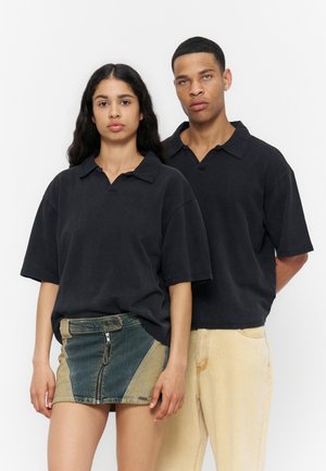 Młoda kobieta w spódnicy z denimu patchworkowego i czarnej koszulce polo stoi obok młodego mężczyzny w beżowych spodniach i dopasowanej czarnej koszulce polo na białym tle.