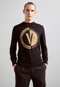 Zwarte gebreide trui met een groot cirkelvormig logo in goud en de tekst "VERSACE JEANS COUTURE", ronde halslijn en geribde manchetten en zoom.