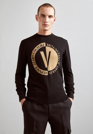 Maglione nero a maglia con un grande logo circolare dorato e la scritta "VERSACE JEANS COUTURE", scollatura a girocollo, e polsini e orlo a coste.