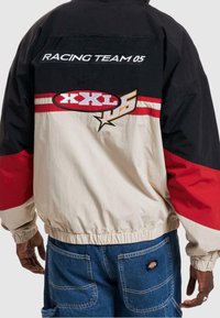 Pestrá větrovka má černý horní díl, béžový střed se červenými akcenty a vyšívané grafiky včetně nápisů "XXL 05" a "RACING TEAM 05".