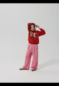 Girl wearing a red sweatshirt with "BC" lettering, pink wide pants, and striped shoes, posing with one arm raised and the other bent.