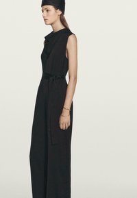 Zwarte mouwloze jumpsuit met een gedrapeerde halslijn en een tailleband. De stof lijkt glad en licht van gewicht, wat zorgt voor een ontspannen pasvorm.