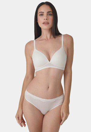 Frau mit langen dunklen Haaren, die einen weißen BH mit dünnen Trägern und passende Spitzenhöschen trägt, stehend vor einem hellen Hintergrund.