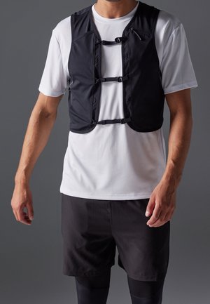 Mand iført hvid kortærmet skjorte og sort vest med tre frontlukninger, kombineret med sorte shorts og leggings.
