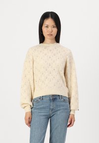 Maglione lavorato a maglia color crema con un motivo a diamante testurizzato, maniche a palloncino e orlo a coste. Abbinato a jeans azzurri chiari.
