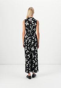 Salopetă neagră cu model floral alb, cu design fără mâneci, talie elastică și deschidere în formă de cheie pe spate. Pantofi plat vizibili.