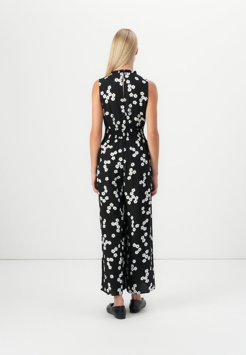 Schwarzer Overall mit weißem Blumenmuster, ärmelloses Design, elastischer Taillenbund und Schlüssellochöffnung hinten. Flache Schuhe sichtbar.