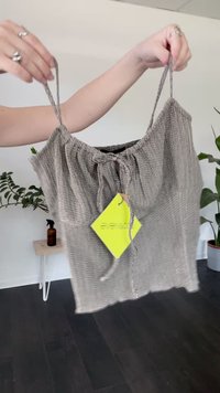 Mani che tengono un top corto grigio a spalla sottile con un'etichetta gialla, piante d'appartamento e un tavolo di legno sullo sfondo.