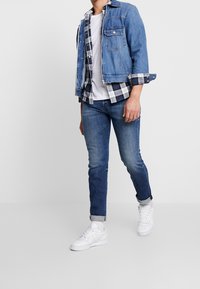 Veste en denim avec deux poches avant, superposée à une chemise à carreaux. Jean bleu et baskets blanches complètent la tenue.