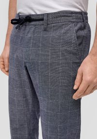 Grijze geruite broek met een elastische tailleband en aantrektouw. De stof is gestructureerd, lichtgewicht en heeft een subtiel ruitpatroon.