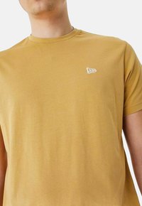 T-shirt à manches courtes et col rond en jaune moutarde, avec un petit logo brodé blanc sur la poitrine gauche. Tissu en coton doux.