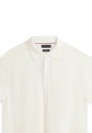 Witte, gebreide polo met korte mouwen, knoopsluiting aan de voorkant en zachte stoffen kraag op een witte achtergrond.