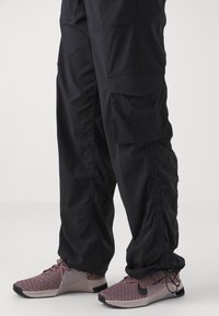 Jambes inférieures portant un pantalon cargo noir avec des cordons de serrage aux chevilles et des baskets Nike mauves avec des semelles blanches.