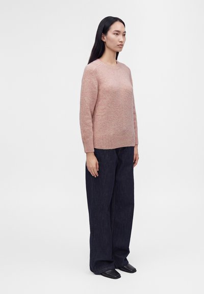 Jeune femme debout portant un pull rose clair, un pantalon large foncé et des chaussures noires sur un fond clair uni.